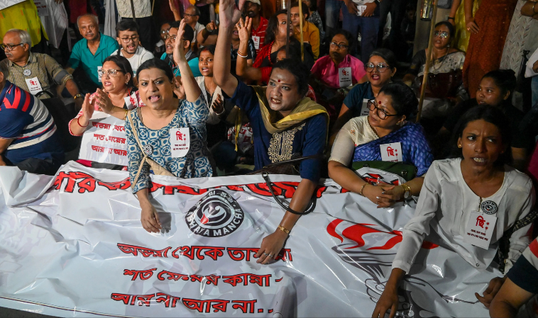 A group of protesters, including women and members of the transgender community, are gathered holding a large banner during a demonstration. Many of them have expressive faces and are raising their hands, shouting slogans. The banner has Bengali text and 